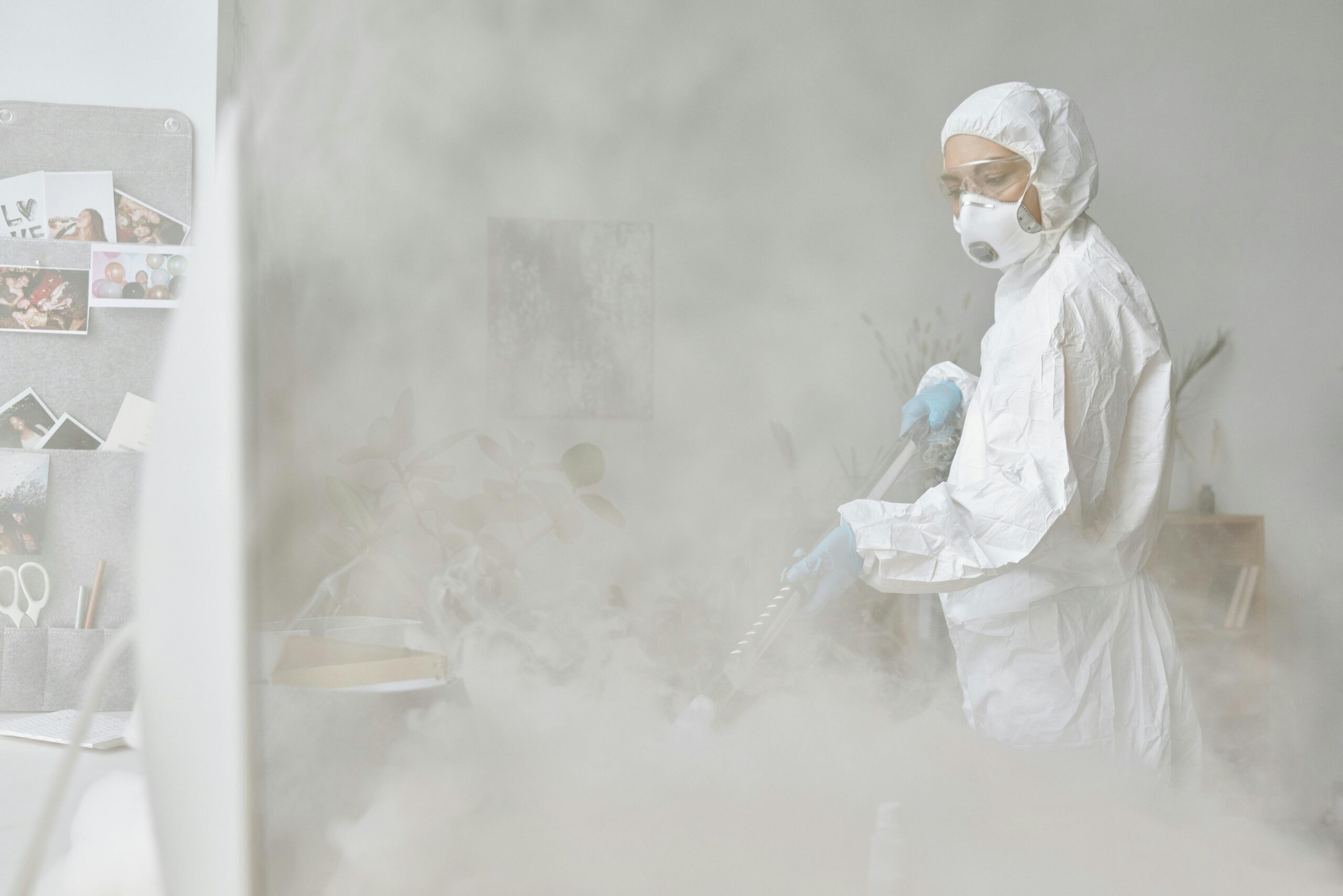 A woman in a protective suit disinfects an indoor space with fogging equipment.